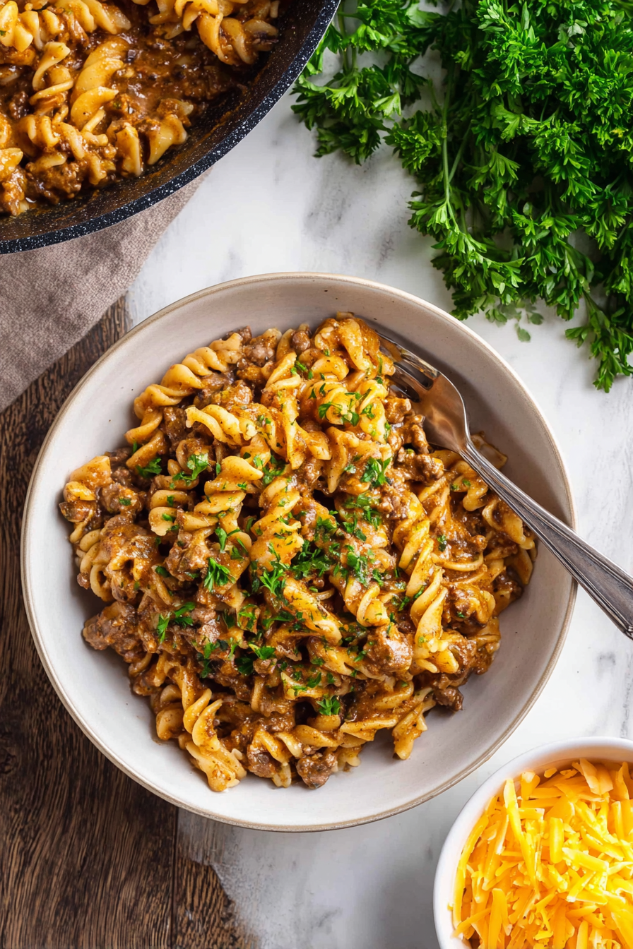 A black pan filled with creamy pasta made of curly rotini noodles mixed with a thick brown and orange sauce, with visible small bits of plant-based meat and sprinkled with fresh green parsley on top. The pan sits on a black and white striped cloth on a dark wood surface. Near the pan is a small white bowl of shredded yellow cheddar cheese and uncooked curly pasta scattered around. In the top left corner, there is a package of plant-based meat with a reddish color. The background shows some fresh green herbs on the right side. Photo taken with an iphone --ar 2:3 --v 7
