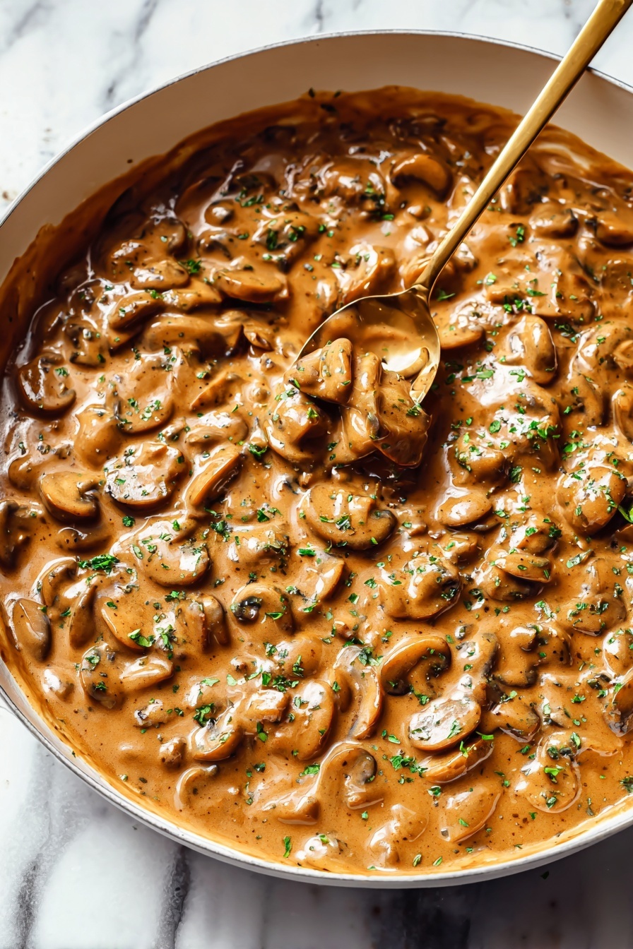 The image shows a thick mushroom sauce in a white deep pan with a smooth, glossy texture. The sauce is a warm brown color with many sliced mushrooms layered evenly throughout. Small green herb pieces are sprinkled on top, adding a fresh touch. A gold spoon is partly dipped into the sauce, lifting some mushrooms and sauce. The pan sits on a white marbled surface, creating a clean and bright background. photo taken with an iphone --ar 2:3 --v 7