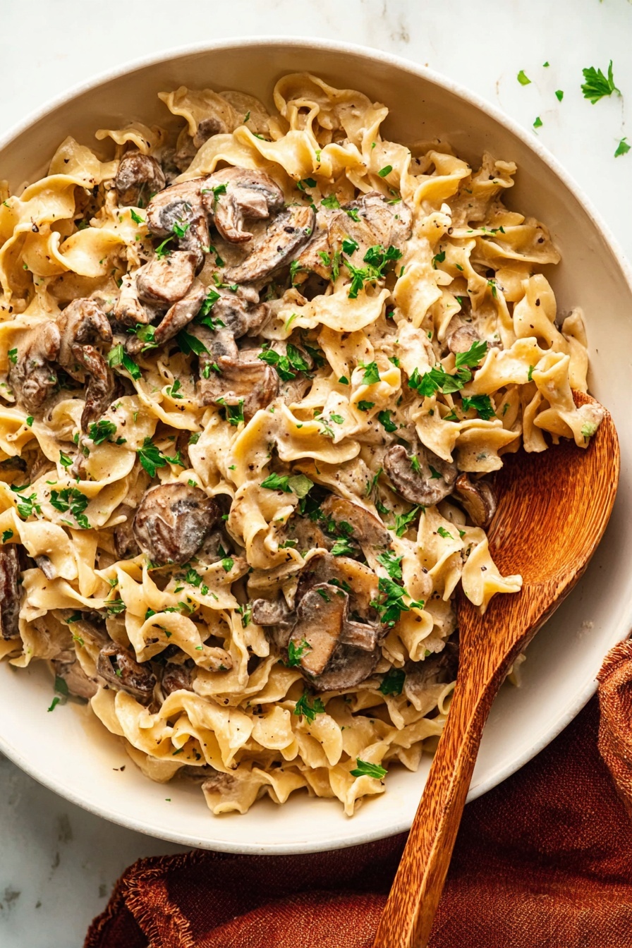 A white bowl filled with creamy pasta shows about three layers: the bottom layer is light beige wide egg noodles with a soft texture; the middle layer is thick slices of browned mushrooms mixed in, some coated in cream; the top layer is chopped green parsley scattered over the dish adding a fresh pop of color. A wooden spoon with a smooth grain pattern is partly buried in the pasta on the right side of the bowl. The bowl rests on a white marbled surface with a folded rust-colored cloth peeking out at the bottom right corner. photo taken with an iphone --ar 2:3 --v 7