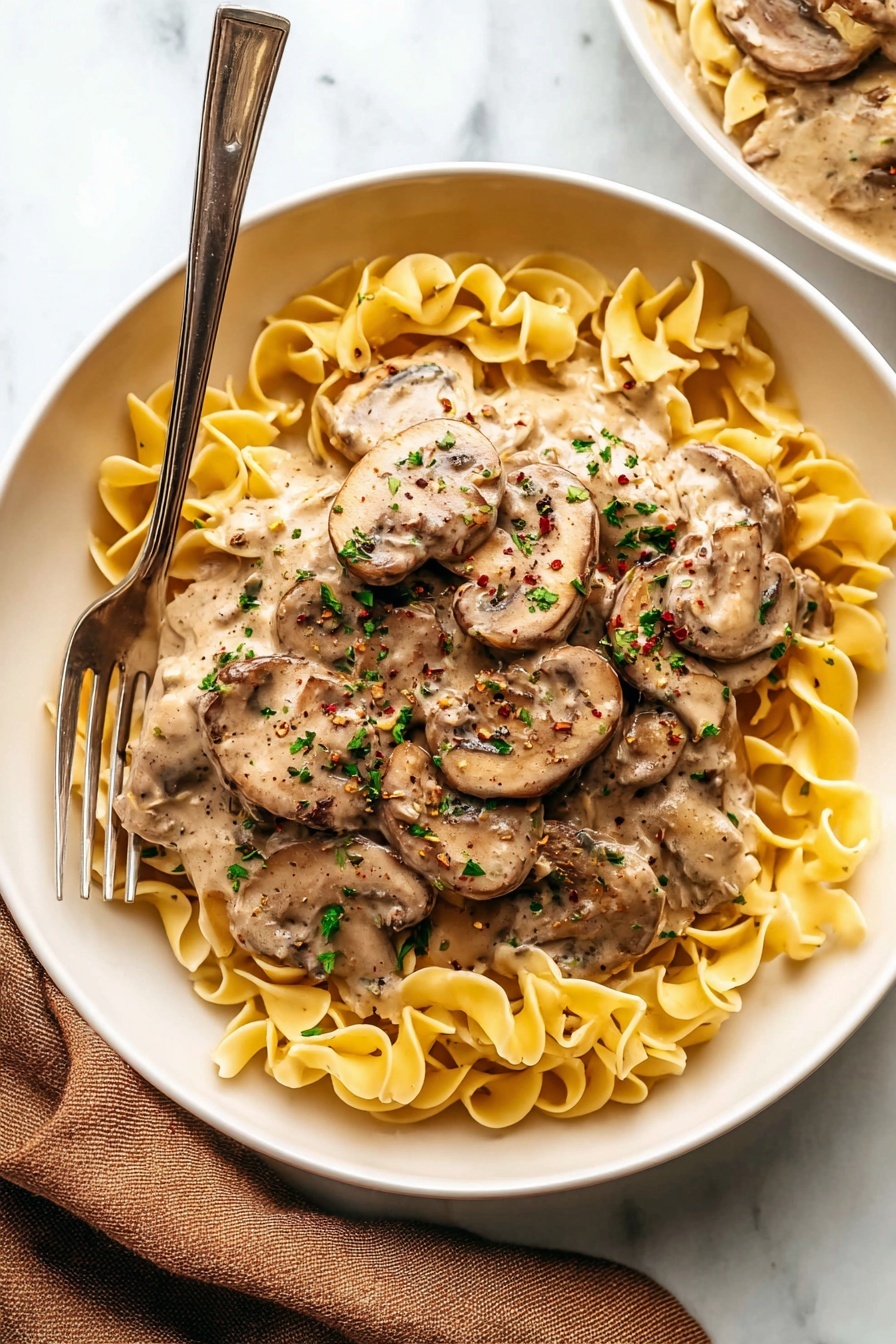 A white bowl holds three layers: the bottom layer is a bed of yellow curly egg noodles with a wavy texture, the middle layer is creamy beige sauce with smooth consistency covering the noodles, and the top layer consists of sliced brown mushrooms mixed into the sauce, sprinkled with small pieces of green herbs and black pepper flakes. A silver fork on the left side leans into the bowl, and a folded brown cloth napkin lies next to the bowl on a white marbled surface. Photo taken with an iphone --ar 2:3 --v 7
