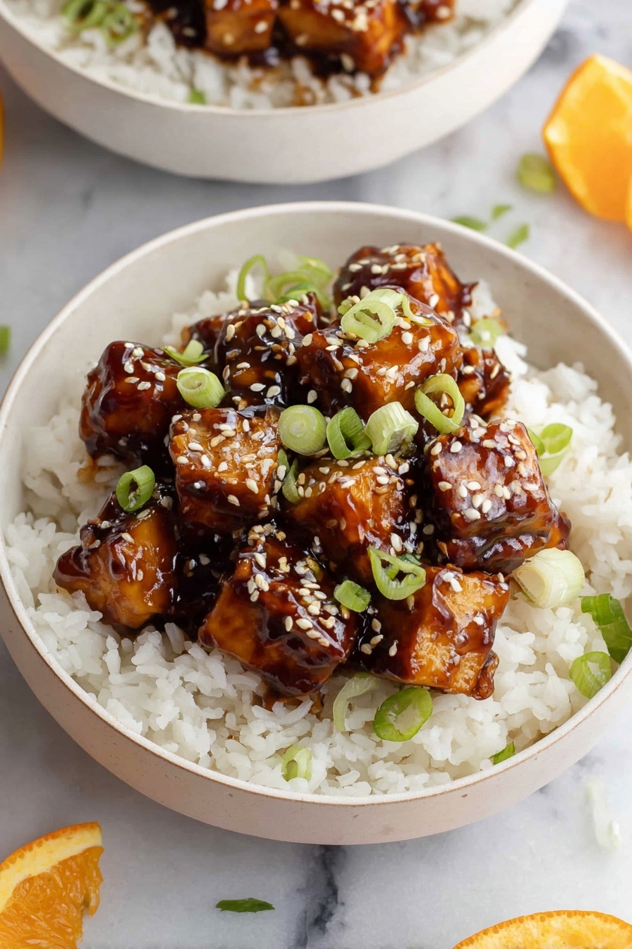 A white bowl filled with a base layer of fluffy white rice topped with a second layer of shiny, sticky dark brown sauce-covered tofu cubes. The tofu is garnished with small light beige sesame seeds and thinly sliced green onions, adding fresh green pops of color. The bowl sits on a white marbled surface, with part of another similar bowl and an orange slice partially visible in the background. Photo taken with an iphone --ar 2:3 --v 7