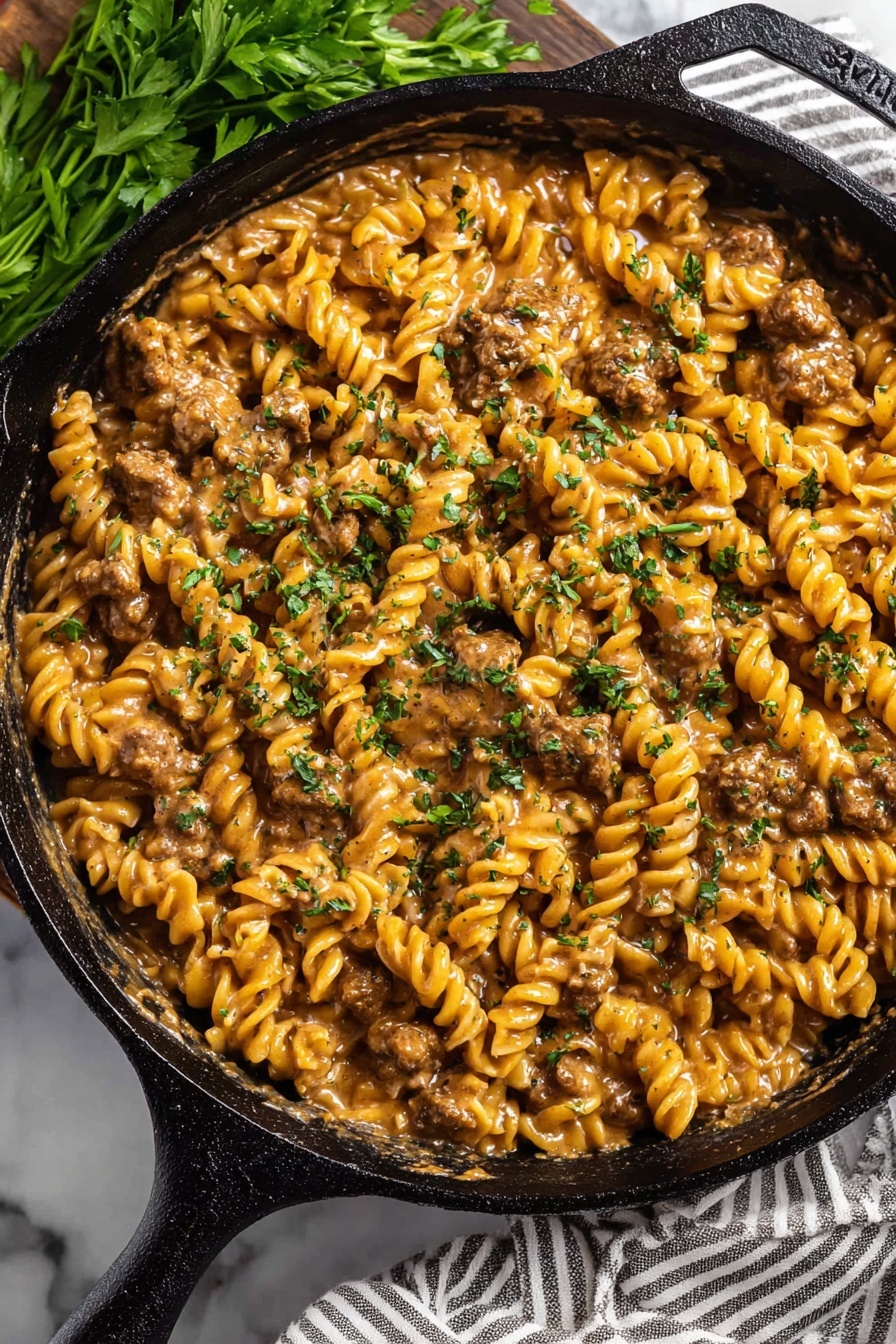 The image shows a close-up of a black cast-iron skillet filled with a creamy pasta dish made of spiral rotini noodles mixed with browned meat pieces, all coated in a rich, light brown sauce. The surface of the dish is sprinkled with finely chopped green herbs that add a fresh, colorful contrast. The skillet sits on a white marbled surface with a striped cloth partially visible underneath, and some fresh green herbs are placed nearby. The food's texture looks thick and creamy with the noodles and meat well blended. Photo taken with an iphone --ar 2:3 --v 7