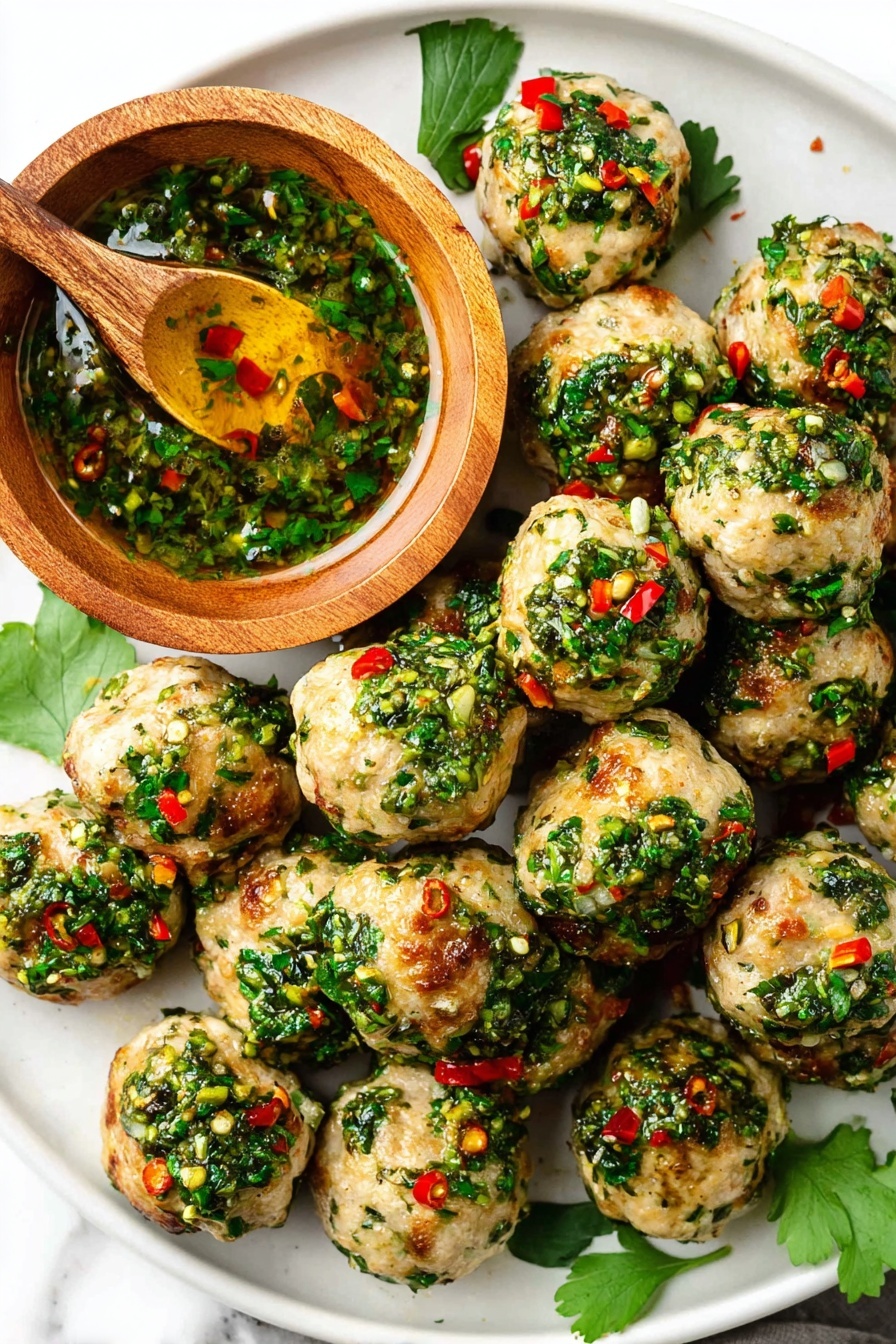 A white plate holds about twenty small round meatballs, each topped with a bright green herb sauce mixed with tiny red chili pieces and bits of garlic, giving a fresh and vibrant look. The meatballs have a light brown, slightly crispy surface with green herbs speckled inside. On the top left of the plate, there is a small wooden bowl filled with extra green herb sauce, showing a shiny, oily texture with visible chopped herbs and red chili pieces. A wooden spoon is inside the bowl, partially covered in the sauce. The plate sits on a white marbled surface, with a few green herb leaves scattered around for decoration. photo taken with an iphone --ar 2:3 --v 7