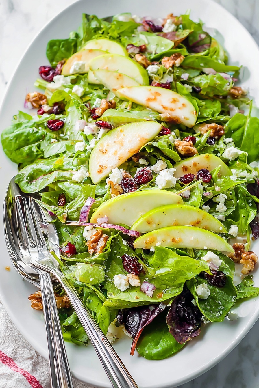 This image shows a fresh salad served on a white oval plate with a white marbled surface background. The salad has three main layers: the bottom layer is a mix of green leafy vegetables with different textures and shades, including dark green spinach leaves and lighter, softer leaves, some with a reddish tint. On top are medium-thick green apple slices placed evenly across the dish, adding a creamy light green color. Scattered throughout are small white crumbles of cheese and reddish dried cranberries, adding spots of white and red. Small pieces of light brown walnuts and finely chopped red onions are sprinkled all over, adding more color and texture. Two silver forks are placed on the left side of the plate, resting on the salad. Photo taken with an iphone --ar 2:3 --v 7