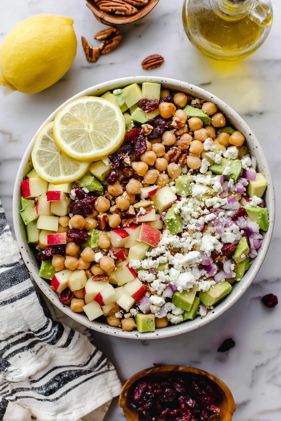 A white bowl filled with a colorful salad layered with chickpeas, diced red apple pieces, avocado chunks, and small red onion pieces mixed evenly. On top, white crumbled cheese is spread out along with scattered dark red dried cranberries and small brown pecan pieces. Two lemon slices rest on the left side inside the bowl. Around the bowl, there is a lemon half on the white marbled surface and a glass bottle of olive oil nearby. A white cloth with black stripes is placed at the bottom left corner of the image. photo taken with an iphone --ar 2:3 --v 7