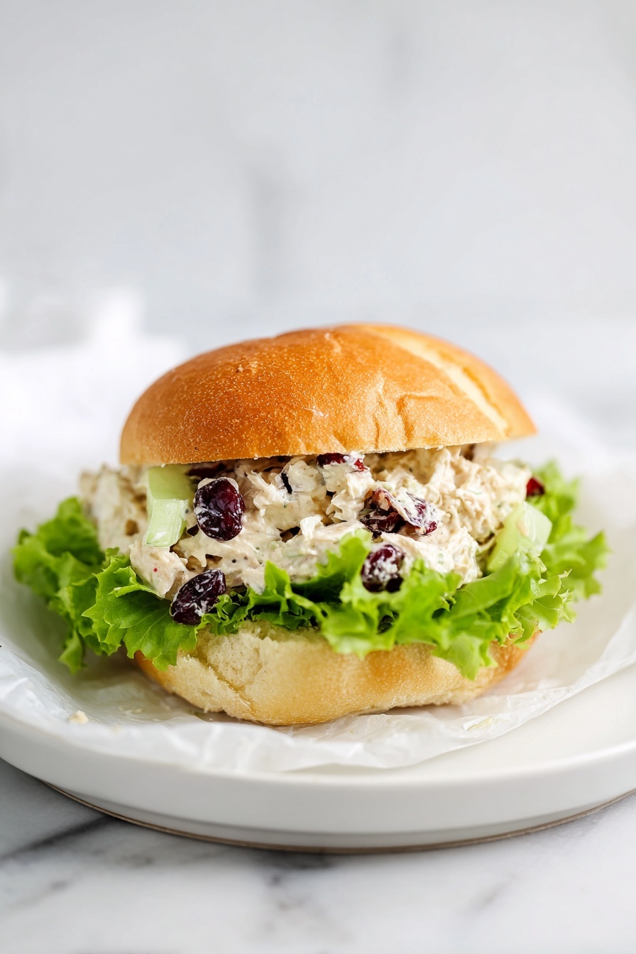 A golden brown sandwich bun with a slightly crispy texture on top holds three layers inside: the bottom layer consists of fresh, ruffled green lettuce leaves peeking out, above it is a thick, creamy white chicken salad mixed with visible small chunks of chopped celery and dark red dried cranberries, and the top bun rests lightly above with a soft, airy texture. The sandwich sits on a white plate lined with white parchment paper, all placed on a white marbled surface. photo taken with an iphone --ar 2:3 --v 7
