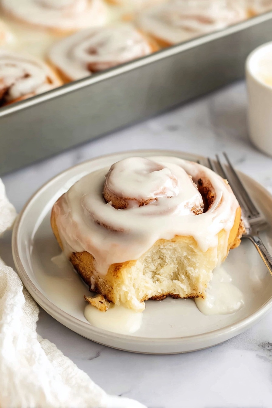 A single cinnamon roll sits on a small white plate, featuring a soft, light brown spiral dough base with visible cinnamon filling swirls. The roll is topped with a thick layer of creamy white icing that drips down the sides, partially melting and pooling slightly at the base. The cinnamon roll has a bite taken out of one side, showing the moist, fluffy interior. In the background, there is a metal baking pan filled with additional cinnamon rolls covered in the same white icing. The scene is set on a white marbled surface with part of a white cloth and a fork visible near the plate. Photo taken with an iphone --ar 2:3 --v 7