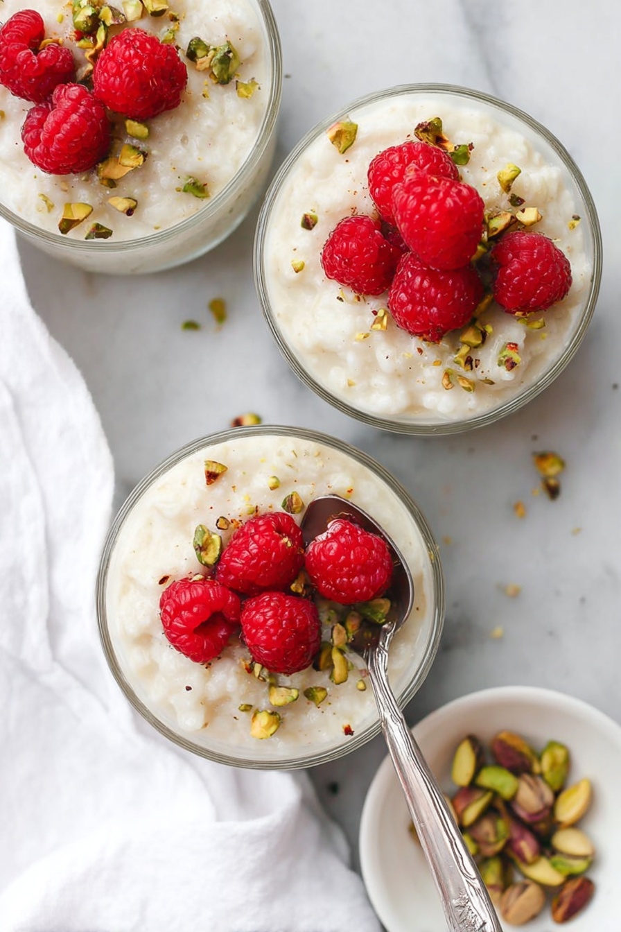 Three clear glass bowls each hold creamy white rice pudding with a soft, smooth texture. On top of each bowl, there are several bright red fresh raspberries clustered together, creating a nice contrast with the white pudding. Scattered around the raspberries and on the pudding are small pieces of chopped greenish-brown pistachios adding texture and color. A silver spoon is placed inside the bowl in the front, resting on the pudding. The bowls sit on a white marbled surface with a white cloth napkin nearby and a small white plate with pistachios visible at the edge. Photo taken with an iphone --ar 2:3 --v 7