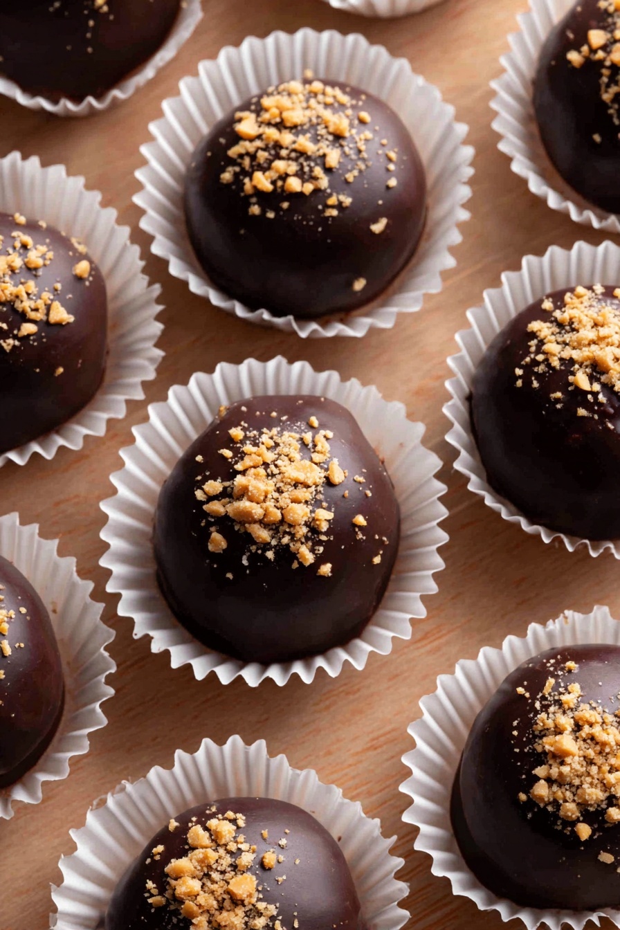The image shows several round chocolate truffles, each placed in a white paper cup with ridged edges. The truffles are dark brown with a smooth, glossy surface and are topped with small, light tan nut pieces crushed finely and scattered in an uneven layer. The white paper cups are evenly spaced on a wooden surface that has a natural, textured look. The truffles are arranged in neat rows, filling the frame with line and repetition. photo taken with an iphone --ar 2:3 --v 7